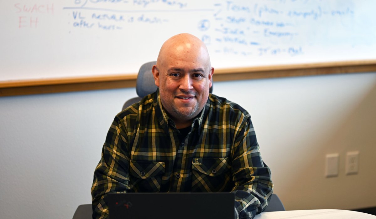 Rueben sits in a rolling chair with a white board with notes behind him. He is smiling at the camera and wearing a black and gold flannel shirt.